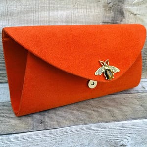 May include: An orange clutch bag with a flap closure. The bag has a gold-coloured bee embellishment and a magnetic snap. The clutch is made of a soft, felt-like material.