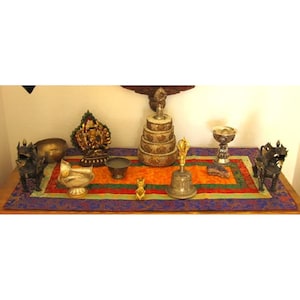 May include: A display of religious and decorative objects, such as statues, bowls, a bell, and a tiered structure, presented on a colourful rectangular mat. The items are made from diverse materials, including metal and wood, and are arranged on a wooden surface.