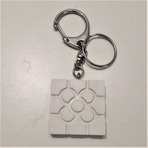 Può includere: Portachiavi in ceramica bianca con un design geometrico. Il portachiavi ha un anello in metallo argentato e una chiusura in metallo argentato.