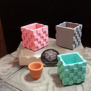 Peut inclure: Trois petits pots carrés en béton avec un motif à carreaux. Les pots sont en rose, gris et turquoise. Un petit pot en terre cuite est également présent sur l'image.