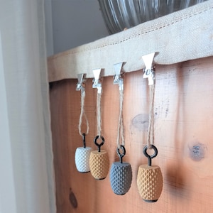 May include: Four decorative table clips with fabric tassels hanging from silver clips. The clips are attached to a beige fabric tablecloth. The tassels are topped with round, textured, decorative balls in white, beige, grey, and brown.