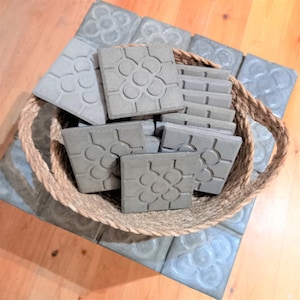 Può includere: Un cesto intrecciato pieno di piastrelle di ceramica grigie con un motivo floreale. Le piastrelle sono disposte in modo casuale e sono tutte della stessa dimensione.