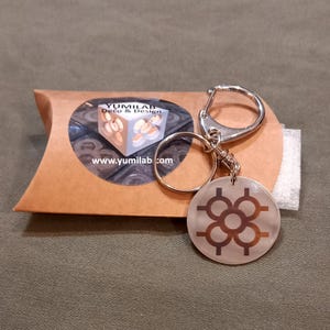 Può includere: Un portachiavi argentato con un ciondolo rotondo argentato che presenta un design geometrico. Il ciondolo ha un motivo metallico marrone. Il portachiavi è confezionato in una busta di carta marrone con un'etichetta bianca che recita "Yumilab Deco & Design" e "www.yumilab.com".