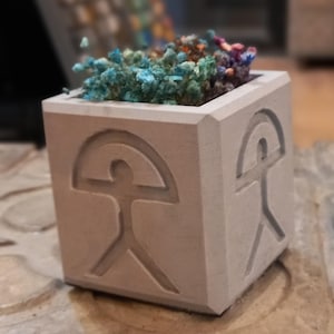Può includere: Un piccolo vaso quadrato in cemento grigio con un design a figura umana scolpito su ogni lato. Il vaso è pieno di fiori secchi blu e viola.