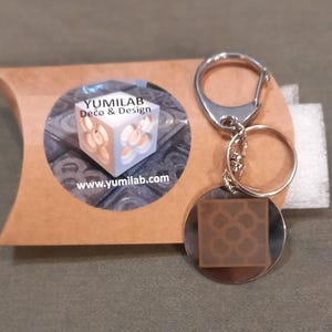 Può includere: Un portachiavi argentato con un ciondolo rotondo argentato che presenta un design geometrico marrone. Il ciondolo ha il testo "YUMILAB Deco & Design" e "www.yumilab.com" su di esso.