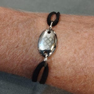 Può includere: Un braccialetto elastico nero con un ciondolo ovale argentato con un design geometrico.