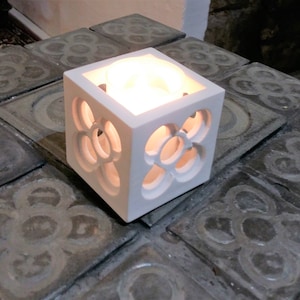 May include: White ceramic candle holder with a decorative cutout design. The holder is cube-shaped and has a small candle inside.