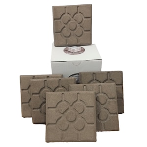 Può includere: Un set di piastrelle decorative quadrate marroni con un motivo floreale intagliato. Le piastrelle sono disposte attorno a una scatola bianca con un logo. Le piastrelle sono realizzate in un materiale strutturato.