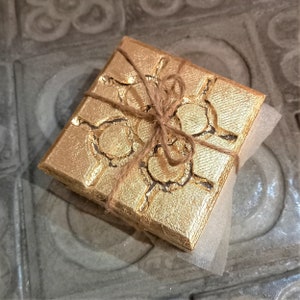 Può includere: Una scatola quadrata color oro con una superficie strutturata e motivi circolari. La scatola è legata con spago e poggia su un pezzo di materiale da imballaggio bianco. Probabilmente si tratta di una confezione regalo o di un oggetto decorativo.