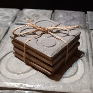 Può includere: Set di quattro sottobicchieri in ceramica grigia con un motivo circolare. I sottobicchieri sono legati insieme con uno spago.