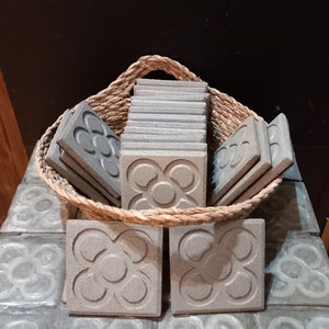 May include: A collection of square, gray concrete tiles with a floral design, displayed in a woven basket. The tiles are stacked and arranged, showcasing their textured surfaces and intricate patterns. The basket sits on a surface with similar tiles.