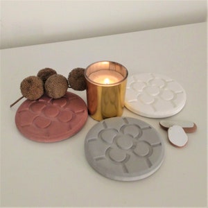 Può includere: Tre sottobicchieri rotondi con un design geometrico. I sottobicchieri sono in tonalità di grigio, bianco e marrone. Un portacandele dorato con una candela accesa è al centro dell'immagine.