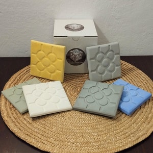 Può includere: Sei sottobicchieri quadrati in ceramica con un design geometrico in rilievo. I sottobicchieri sono di vari colori: giallo, bianco, grigio, grigio chiaro e blu. I sottobicchieri sono disposti su un tappetino di paglia intrecciata. Una scatola bianca con il testo "Yumilab" è sullo sfondo.
