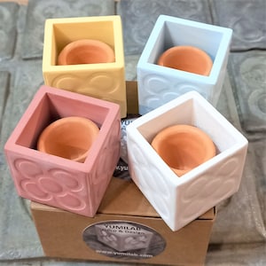 Op de afbeelding: Vier kleine, vierkante potten in geel, lichtblauw, roze en wit. Elke pot heeft een kleine, ronde terracotta pot erin. De potten hebben een reliëf, bloemmotief.