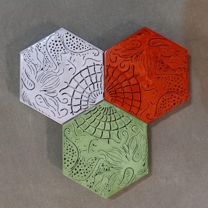 Könnte beinhalten: Drei sechseckige Keramikfliesen mit komplizierten Designs. Die Fliesen sind weiß, rot und grün mit schwarzen Umrissen.