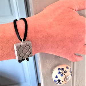 Può includere: Un ciondolo quadrato grigio con un motivo geometrico è appeso a un braccialetto con cordino di velluto nero. Il ciondolo è attaccato al braccialetto con un anello di metallo color argento. È visibile anche un pomello floreale bianco e blu.