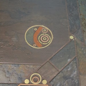 May include: A close-up of a slate floor with a geometric design. The design features a circle with concentric circles and a curved line in brown, yellow, and green. The floor is made of different colored slate tiles.