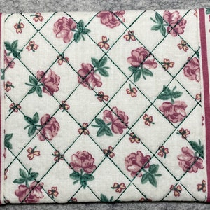 May include: A white quilted fabric with a pink and green floral pattern. The fabric has a diamond-shaped quilting pattern and a red border.