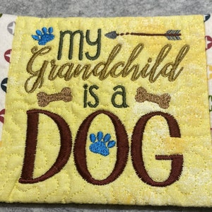 May include: A yellow quilted coaster with the text "my grandchild is a DOG" embroidered in brown and green thread. The coaster has a blue paw print and two brown bone designs. The edges of the coaster are trimmed with a white fabric with red, yellow, and blue stars.
