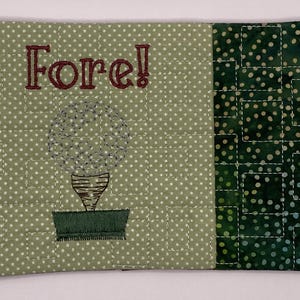 May include: A quilted golf-themed coaster with a green and white polka dot pattern. The word "Fore!" is embroidered in maroon thread above a golf ball design. The right side features a green geometric pattern.