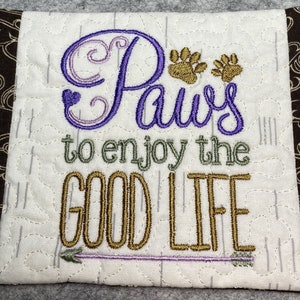 May include: A white quilted coaster with brown fabric trim. The coaster features embroidered text that reads "Paws to enjoy the good life" with a purple heart, paw prints, and an arrow.