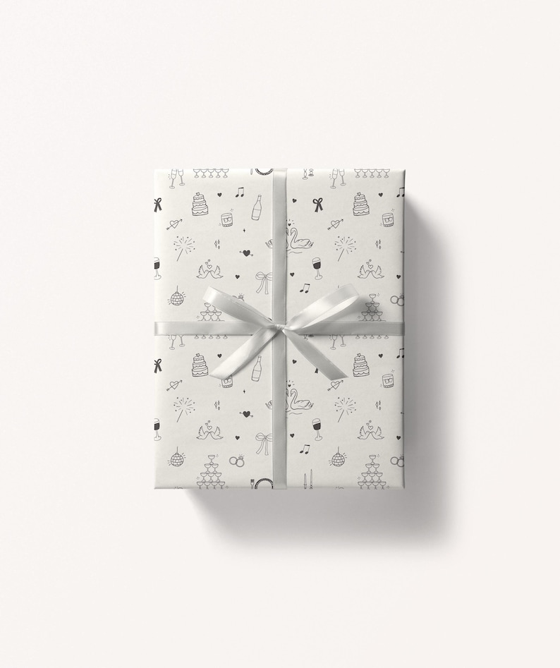 May include: A white gift box with a silver ribbon. The box is wrapped in white paper with a black and white pattern of wedding-themed illustrations, including cake, swans, rings, and champagne glasses.