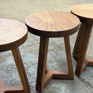 May include: Three wooden stools with round seats and angled legs. The stools are made of a light brown wood with visible grain patterns. The legs are joined at the base, creating a geometric design. The stools are sturdy and suitable for seating.