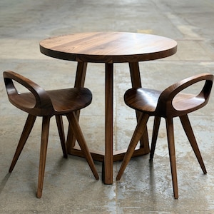 May include: A small, round wooden table with two matching chairs. The table and chairs are made of dark-stained wood. The chairs have a unique design with open backs and angled legs. The table is approximately 76 cm in diameter.