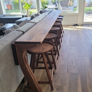 K&ouml;nnte beinhalten: Ein dunkler Holz-Bartresen mit geschwungenem Design und f&uuml;nf passenden Barhockern. Der Tresen ist an einem grauen Sofa befestigt und die Hocker sind in einer Reihe angeordnet.