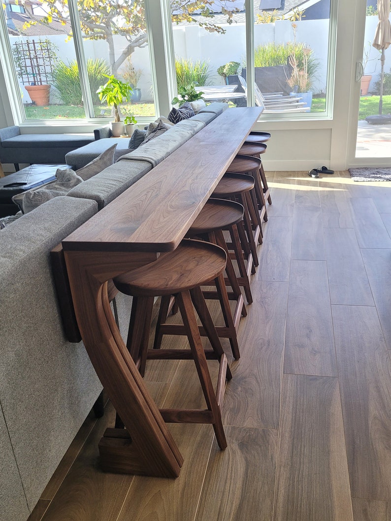 K&ouml;nnte beinhalten: Ein dunkler Holz-Bartresen mit geschwungenem Design und f&uuml;nf passenden Barhockern. Der Tresen ist an einem grauen Sofa befestigt und die Hocker sind in einer Reihe angeordnet.