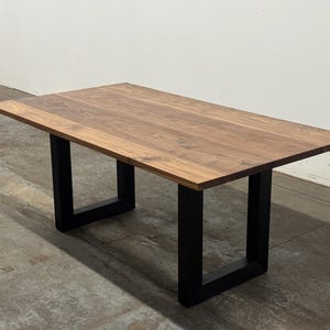 May include: A rectangular wooden table with a natural wood grain finish. The table is supported by black, U-shaped legs. The table's dimensions are approximately 60 inches long and 30 inches wide.