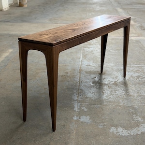 May include: A long, narrow wooden console table with a dark brown finish. The table has a rectangular top and four slender, curved legs. The table is approximately 60 inches long, 12 inches wide, and 30 inches tall.