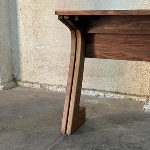 K&ouml;nnte beinhalten: Nahaufnahme eines Tischbeins aus Holz mit einem einzigartigen, geschwungenen Design. Der Tisch ist aus dunkelbraunem Holz gefertigt und hat einen schlichten, modernen Stil.