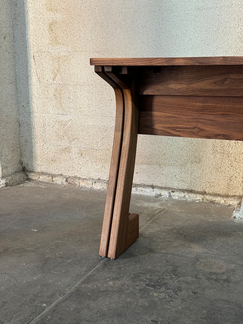 K&ouml;nnte beinhalten: Nahaufnahme eines Tischbeins aus Holz mit einem einzigartigen, geschwungenen Design. Der Tisch ist aus dunkelbraunem Holz gefertigt und hat einen schlichten, modernen Stil.