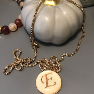 May include: A gold chain necklace with a round pendant featuring the letter "E" in a cursive script. The pendant is white with a gold rim.