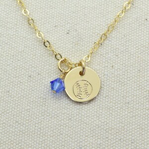 May include: A gold chain necklace with a small gold charm featuring a baseball design. A small blue crystal dangles from the charm.