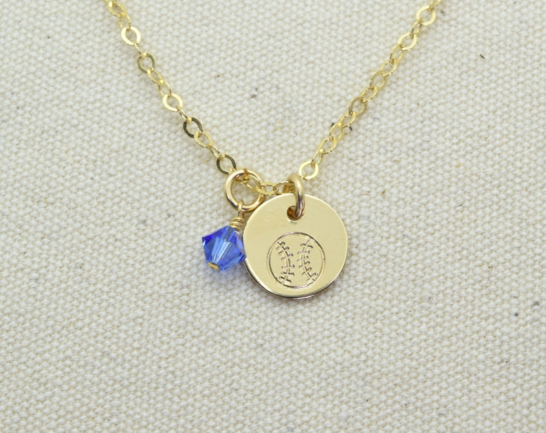 May include: A gold chain necklace with a small gold charm featuring a baseball design. A small blue crystal dangles from the charm.