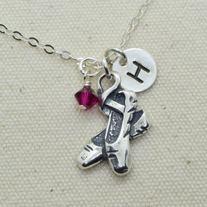 May include: Silver necklace with a charm of a pair of ballet shoes, a small red crystal, and a silver disc with the letter "H" on it.