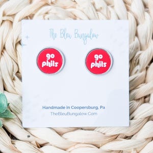 May include: A pair of round stud earrings featuring a bright red background with the white text 'go phils' printed on each. The earrings are attached to a white display card that reads 'The Bleu Bungalow' and 'Handmade in Coopersburg, Pa'.
