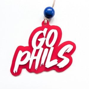 May include: A red and white decorative ornament with the words "GO PHILS" in a bold, layered design. The ornament is suspended by a string and a blue bead.