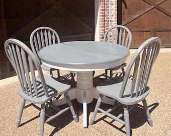 May include: A round, gray and white painted table with four gray painted chairs. The table and chairs are made of wood and have a simple, farmhouse style.