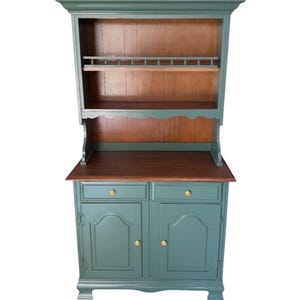 May include: A two-piece hutch in a teal green color with a dark wood countertop and interior. The upper section has a shelf and a rack for displaying items. The lower section has two drawers and two cabinet doors with gold-colored knobs.