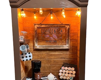 May include: A brown coffee station with a sign that reads "Coffee & Tea Freshly Brewed Served Daily." It includes a coffee maker, mugs, a sugar bowl, and a selection of coffee pods. The station is illuminated by string lights.