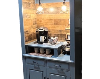 May include: A gray wooden coffee bar cabinet with a glass front, two doors, and two drawers. The cabinet has a built-in countertop with a coffee maker, mugs, and a coffee bar sign. The cabinet has a sign that says "Coffee Bar" above the glass front.