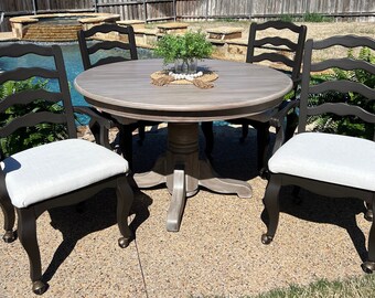 May include: A round wooden dining table with a gray wash finish and four black ladder back chairs with white cushions. The table and chairs are set up on a patio with a pool in the background.