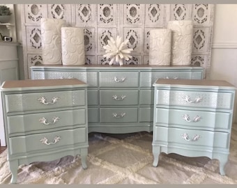 May include: A bedroom furniture set in a soft blue-green color, including a dresser and two nightstands. The set features ornate silver drawer pulls and decorative detailing. The tops of the furniture are a light brown color. Decorative white vases and a white starburst are on top.