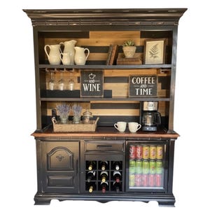 May include: Black wooden hutch with a coffee station and wine storage. The top shelf displays white pitchers, wine glasses, and framed art. The lower section has a wine rack, a mini-fridge with canned drinks, and a coffee maker.