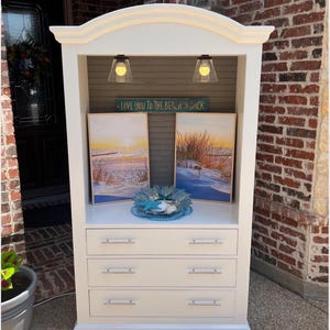 May include: A white cabinet with three drawers and a top shelf, featuring two framed beach landscape paintings and a decorative blue glass bowl with a starfish. The cabinet has a curved top and two small lights. A sign reads "LOVE YOU TO THE BEACH & BACK."