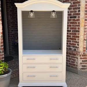 May include: A cream-colored cabinet with three drawers and a display area. The cabinet has a curved top and two small lights. The drawers have silver handles. The cabinet is set against a brick wall.