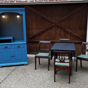 May include: A blue painted hutch with two doors and two drawers, a shelf, and two light bulbs. The hutch is in front of a wooden garage door. There are four chairs and a table with a dark top in front of the garage door.